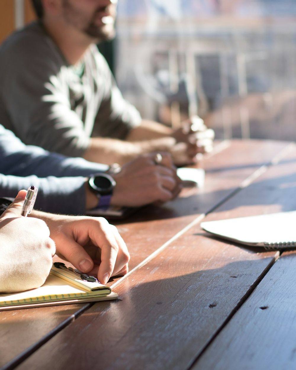 Three people seated at a long timber table, writing notes during a working discussion.