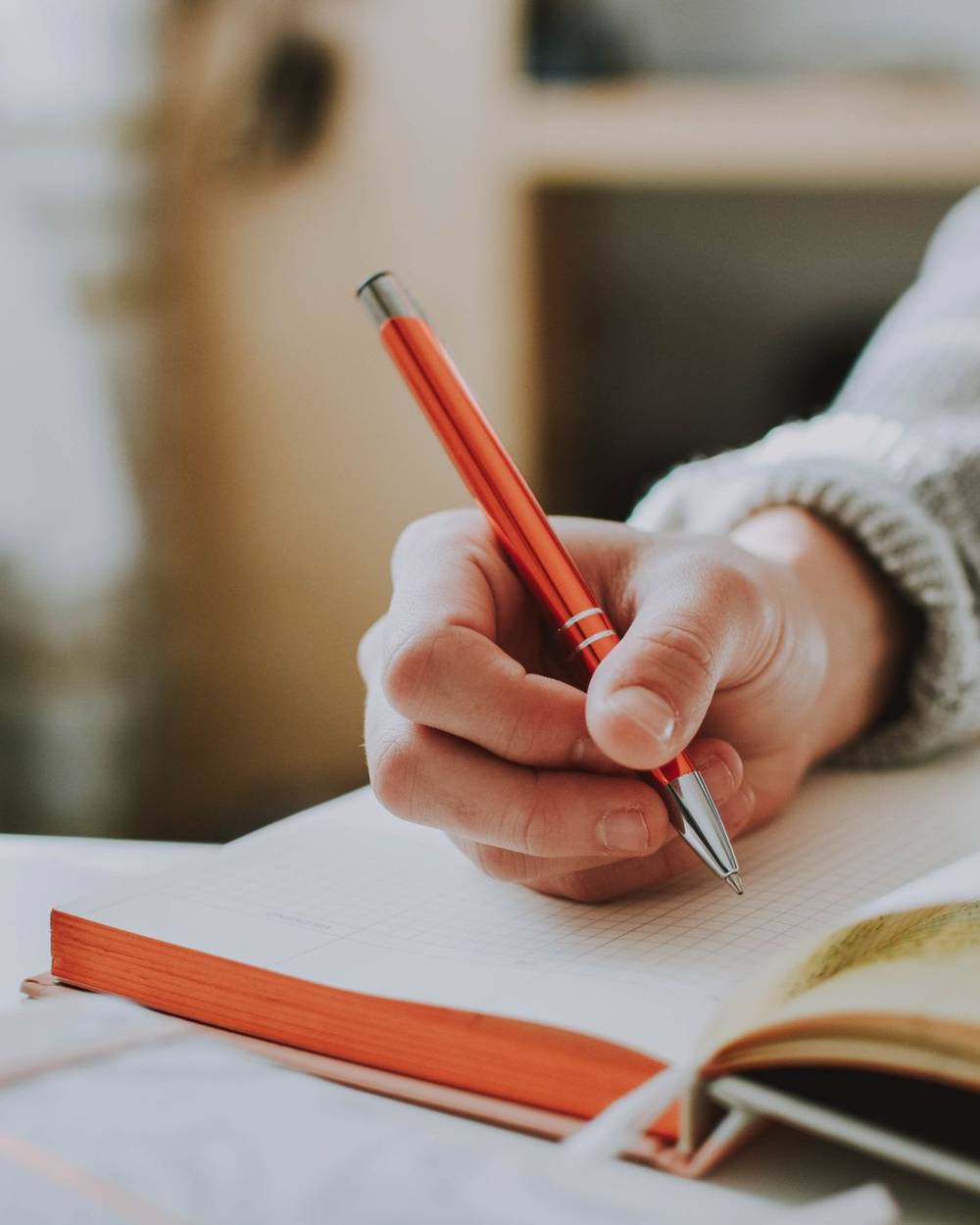 A hand writing notes in a notebook beside an open book, in soft daylight.